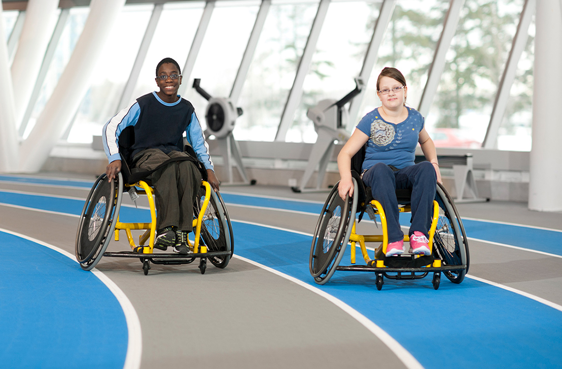 Two children who use wheelchairs racing on a track.