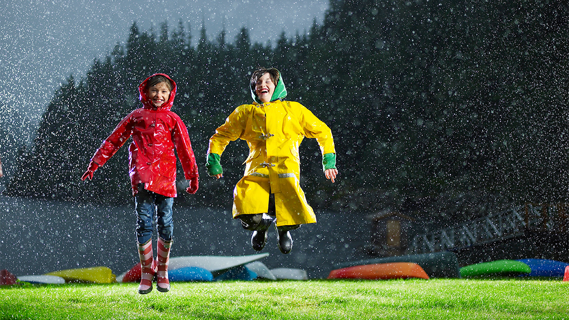 Two children play outside in the rain. They are wearing raincoats and rainboots.