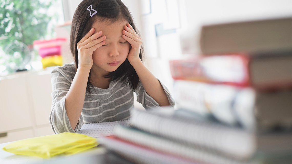 A child is sitting at a desk with a pile of books in front of them. They have their hands on their forehead and appear tired.