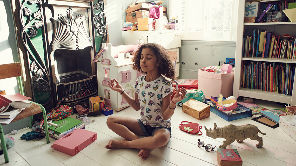 A youth sitting cross-legged on the floor of a bedroom with their eyes closed.