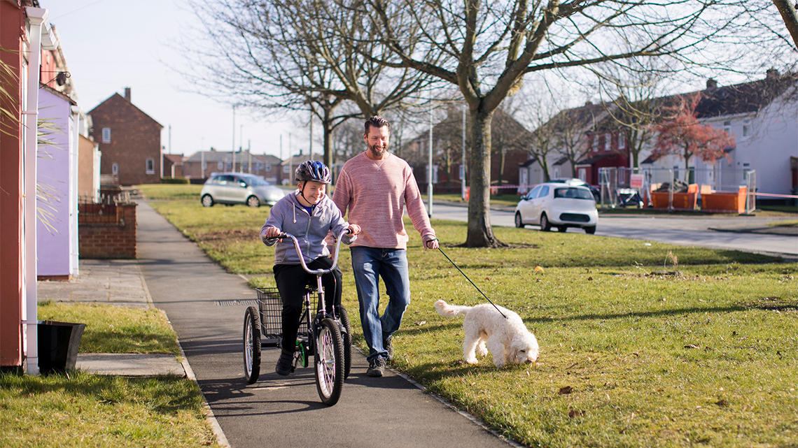 A child is on a bicycle and an adult is walking beside them with a dog on a leash