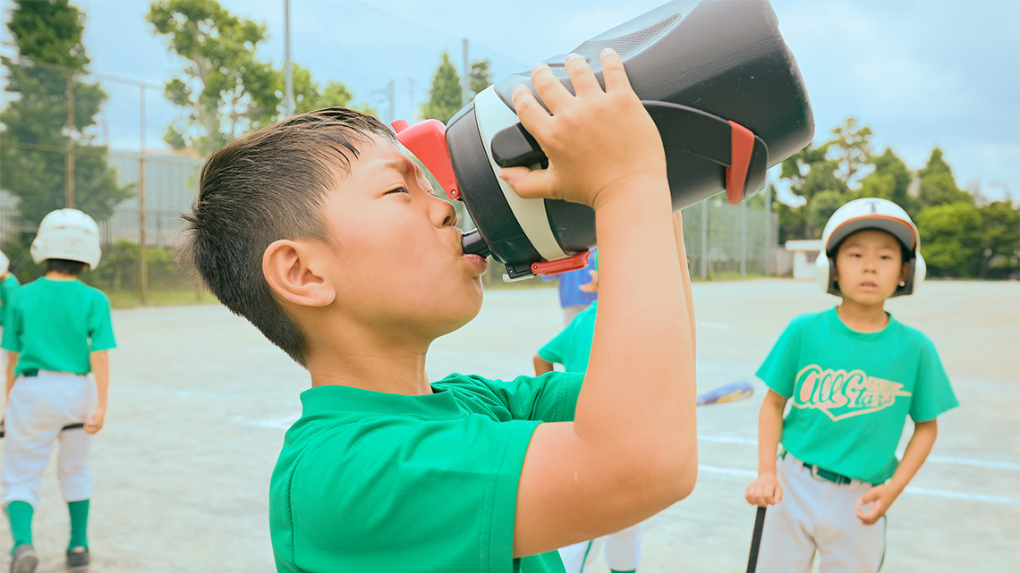 A youth in a baseball uniform drinking from a large water bottle.