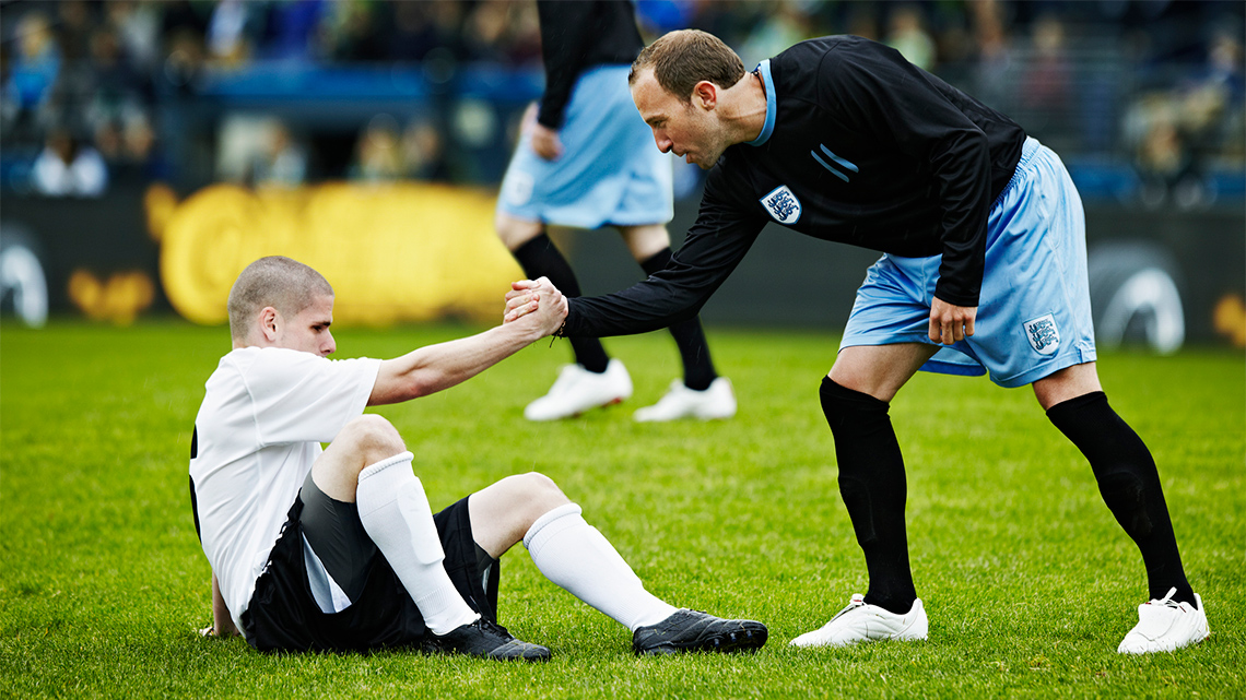 An athlete has fallen on a field. Another athlete on the opposing team is grasping their hand to help them back up.