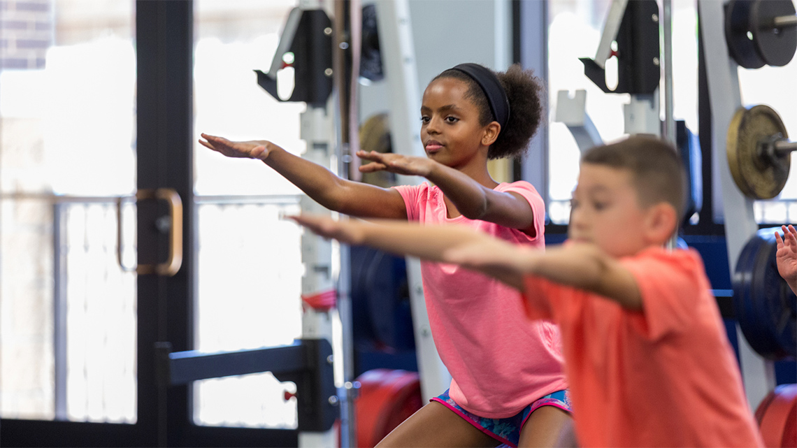 Two children in a gym squatting with their arms raised in front of them.