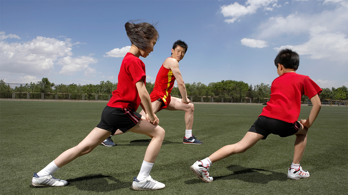 Three people on a field performing lunges by stepping one foot forward, bending the knee, and extending their other leg behind them.