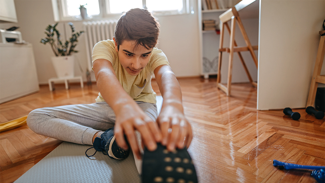 A child sitting on their bedroom floor, stretching out one leg, tucking their other foot against their thigh, and reaching for their toes.