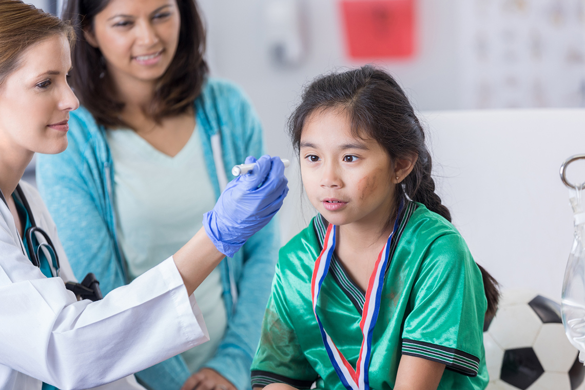 Child with a possible concussion and holding the forehead at the doctor’s office, with their parent.