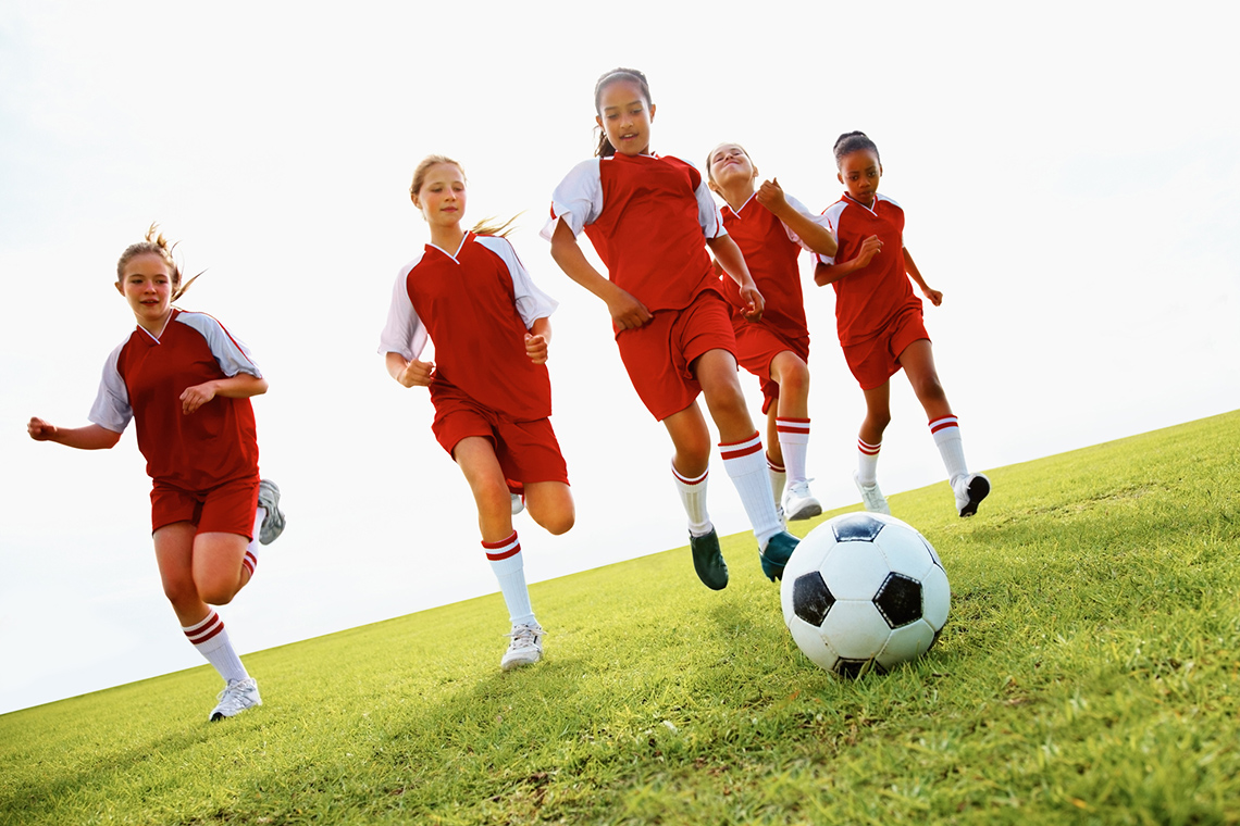 Five girls wearing soccer uniforms run across a grassy field towards you chasing a soccer ball.