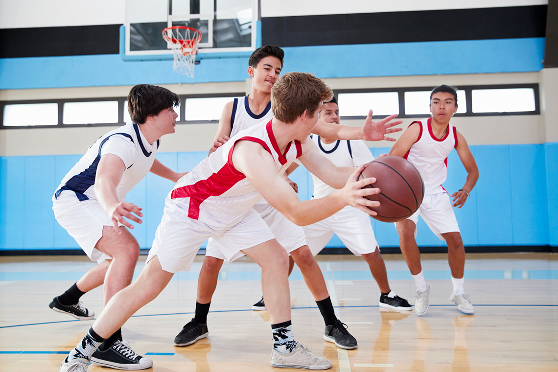 Five boys wearing basketball uniforms play basketball in front of a basketball net on an indoor basketball court.