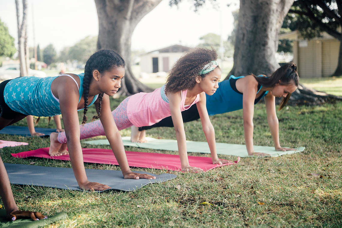 A row of three girls holds themselves up in a push up on exercise mats set up in front of two mature trees in a neighborhood park.