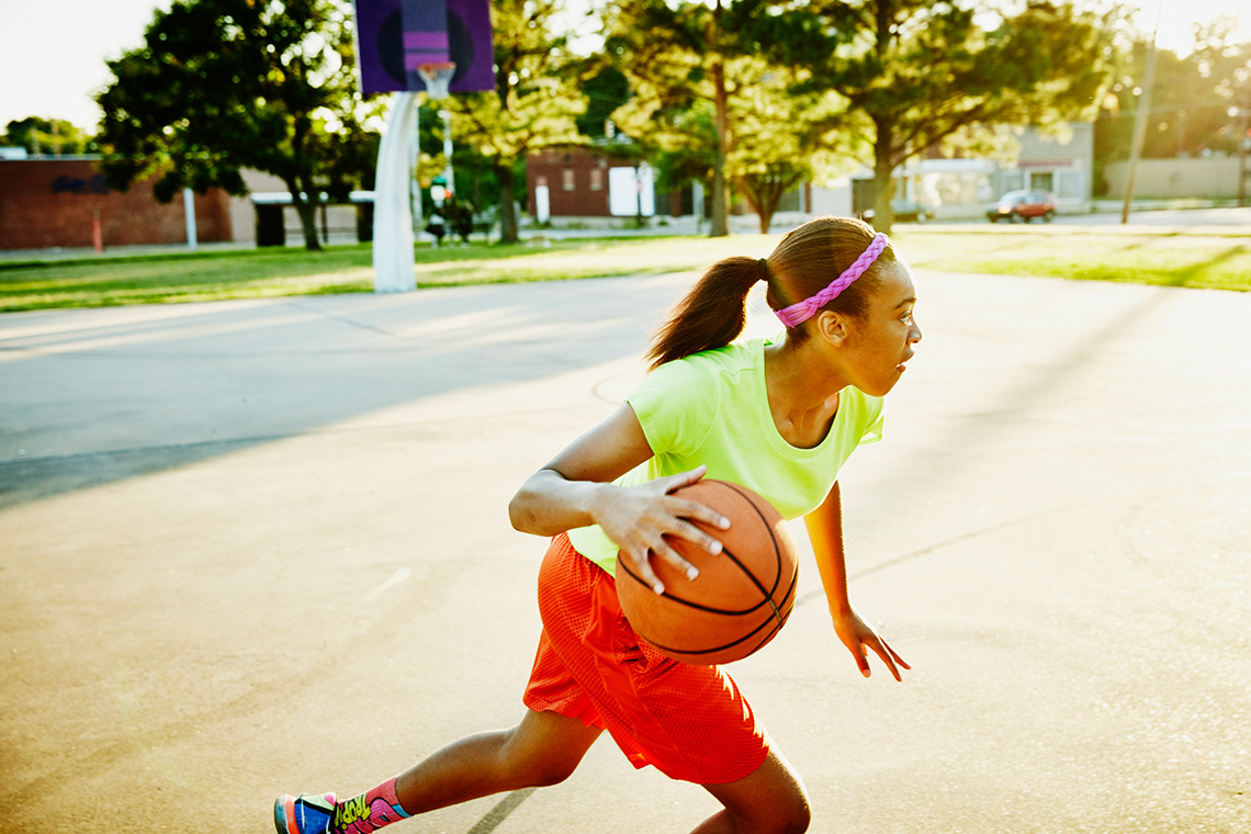 A girl bounces a basketball while moving across a sunny outdoor basketball court.