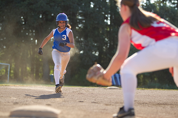 A young person runs on a baseball diamond towards a base guarded by a baseball player wearing a baseball glove outdoors on a sunny day.