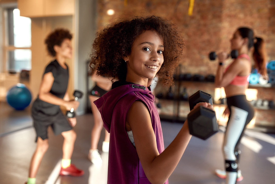 A smiling girl lifts a small barbell weight with her right arm while working out in an indoor gym.