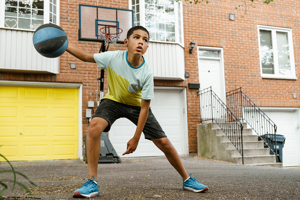 A boy dribbles a basketball outside on pavement in front of some townhouses with a portable basketball net set up between two garage doors.