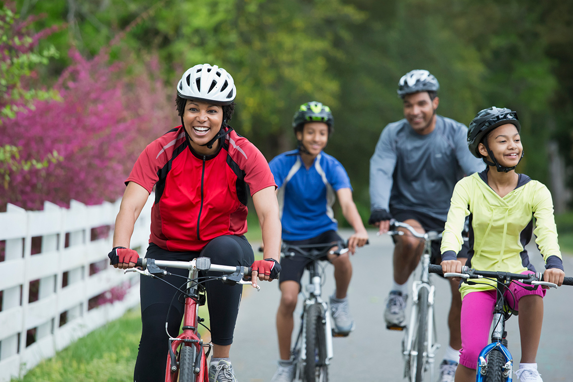 Four people, including a man, a woman, and two children, all wearing helmets, ride their bicycles towards you on the side of a paved road.