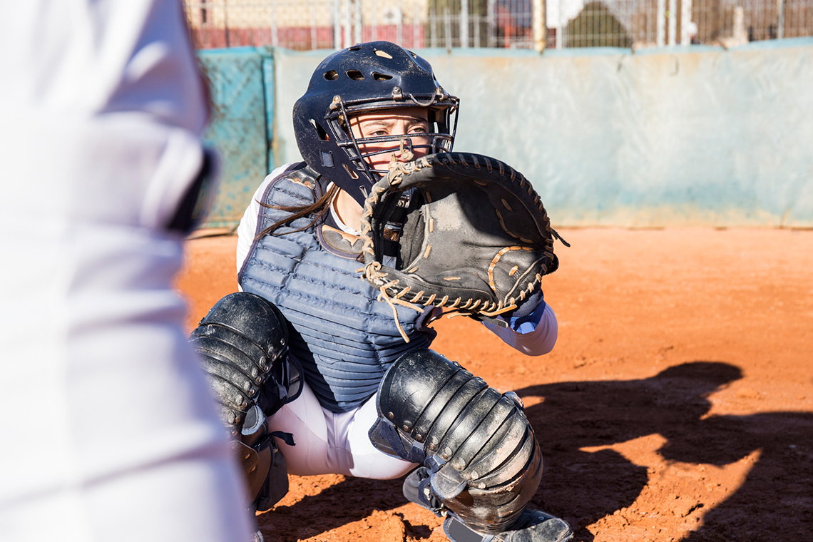 A young baseball catcher, wearing a chest protector, helmet, mask, and knee guards, sits in a crouch and holds up a catcher’s mitt.