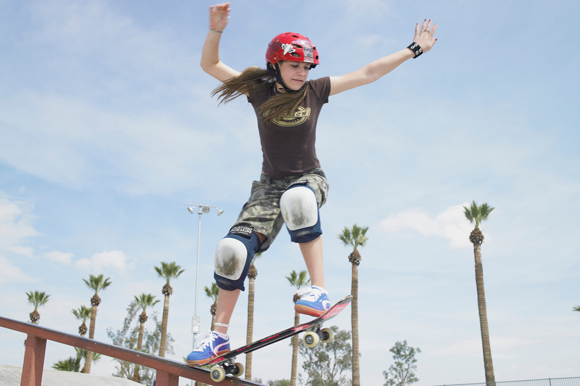 A young person wearing a helmet and knee pads rides their skateboard on its two back wheels down a ramp at an outdoor skateboard park.