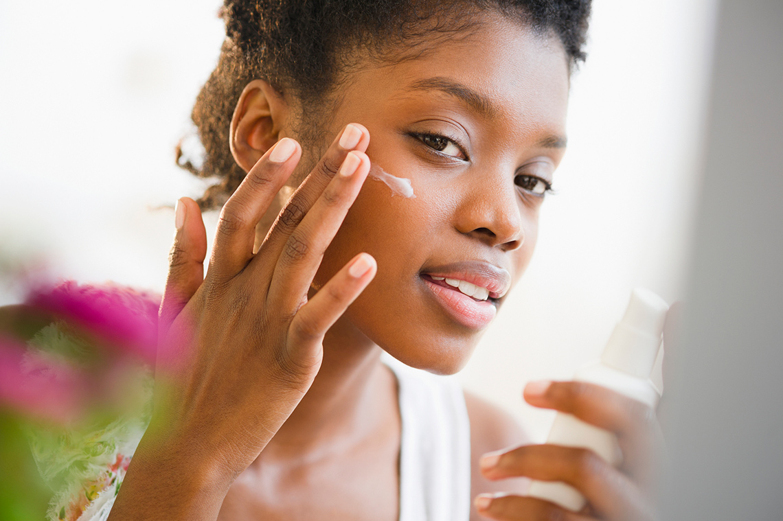 A young person applies sunscreen to their cheek in an indoor bathroom while holding the bottle of sunscreen in their other hand.