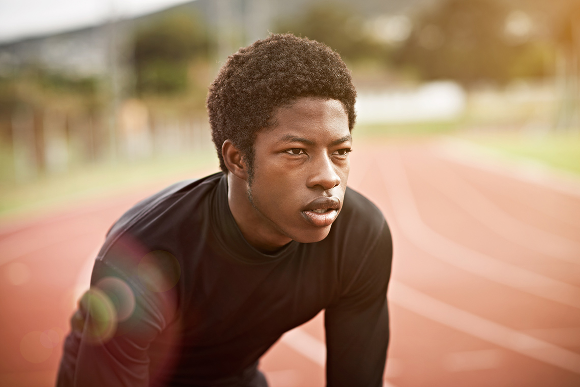 A young person wearing a long-sleeved athletic training shirt bends slightly forward to catch their breath outdoors at a running track.