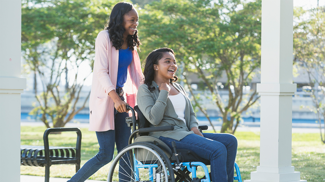On a sunny day, one young person walks along a paved path past a metal bench while pushing a second young person in a wheelchair.