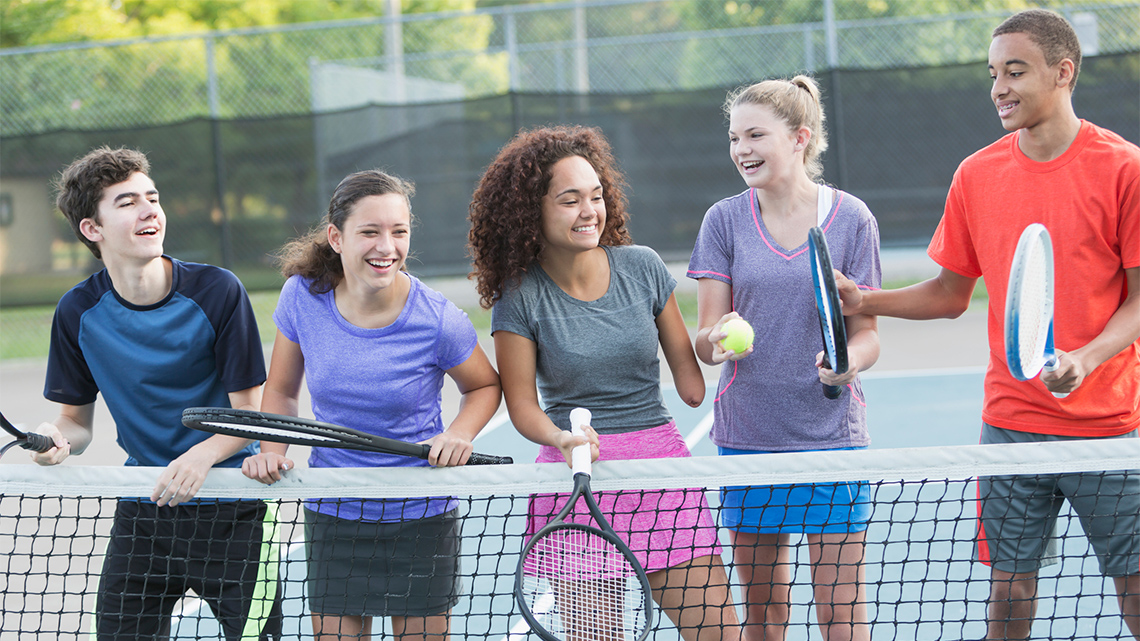 Five young people wearing athletic clothing stand in a row along a net on an outdoor tennis court holding their tennis racquets.