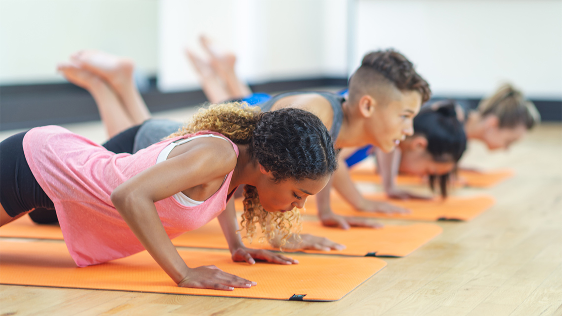 Four young people wearing workout clothes are lined up in a row doing push-ups on gym mats indoors in a gymnasium.