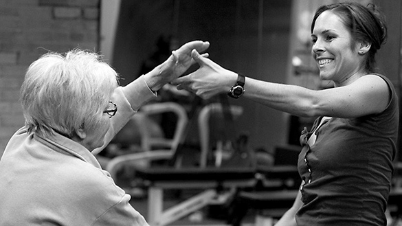 One older adult is about to grasp the hand of a middle-aged adult as they dance indoors in a studio room.