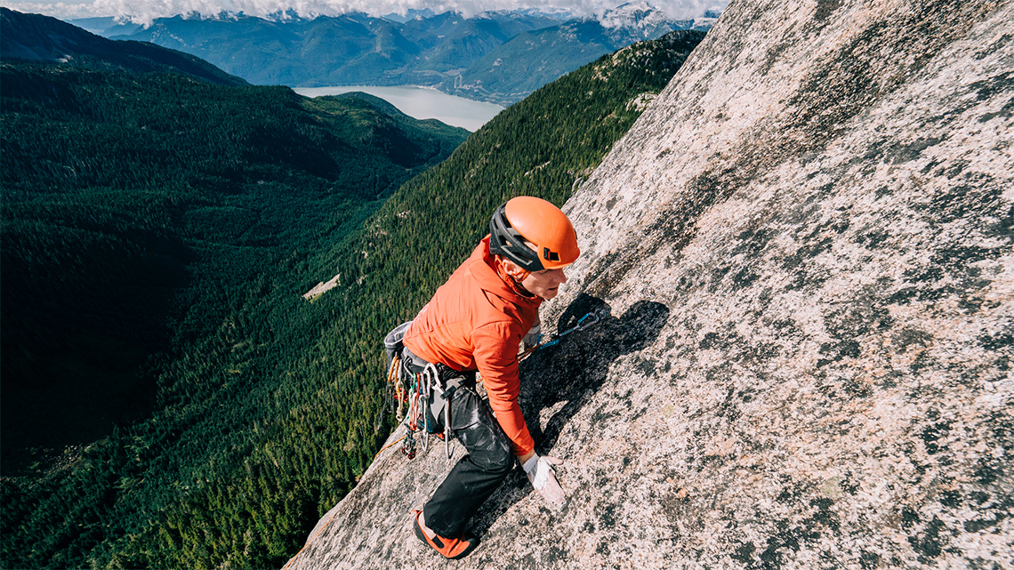 A young person wearing climbing gear, a helmet, and bright clothing carefully climbs up a steep rocky cliff.