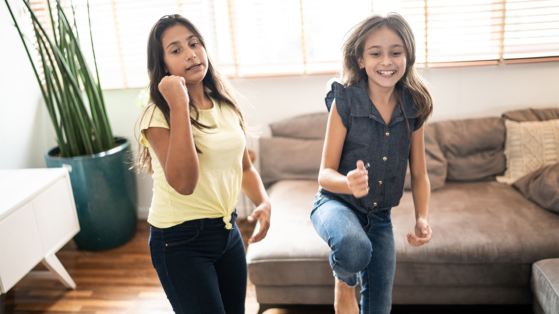 Two young people wearing casual clothing dance indoors to music or to a video in front of a screen in a bright living room.