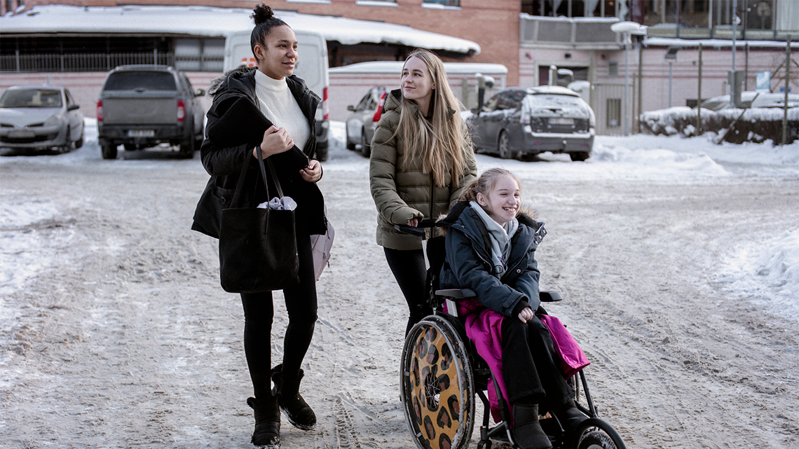 Two young people in winter clothes walk in a snowy parking lot while pushing a third young person, also in winter clothes, in a wheelchair.