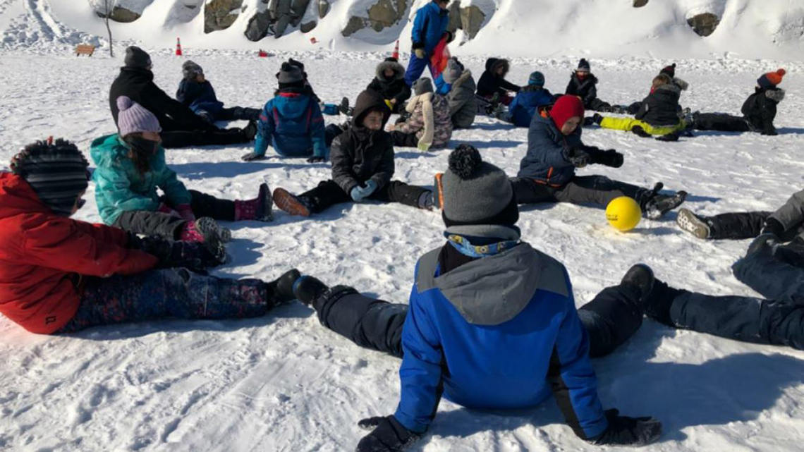 A group of students, wearing snowsuits and winter clothing, sit with their legs spread out on the snow in two circles on a cold sunny day.