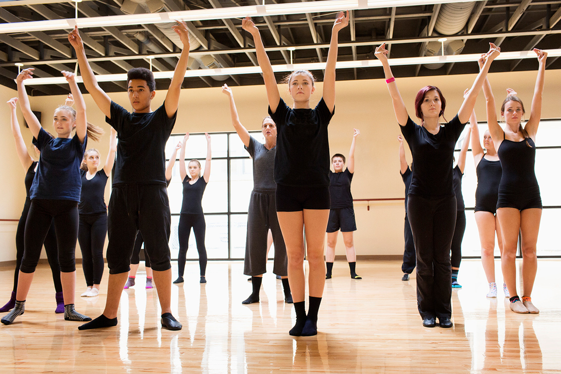 A group of young people stand in an indoor dance studio, their arms raised above their heads, copying movements one of them is performing.