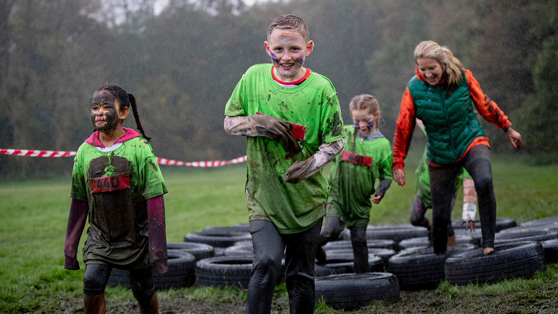 A group of four young people wearing muddy casual clothing walk through a tire obstacle course outdoors on a cloudy and misty day.