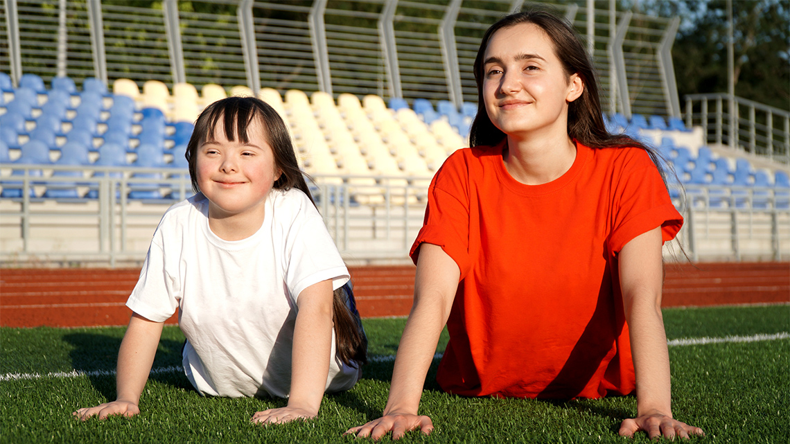 A young child and an older child lie on their fronts outside on the grass in a stadium holding themselves up with their arms in a yoga pose.