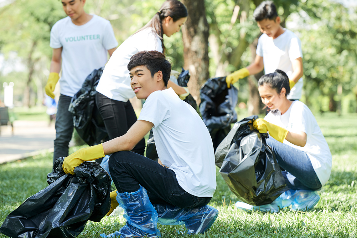 Five young people wearing t-shirts, jeans, and rubber gloves carrying garbage bags pick up trash in an outdoor park on a sunny day.