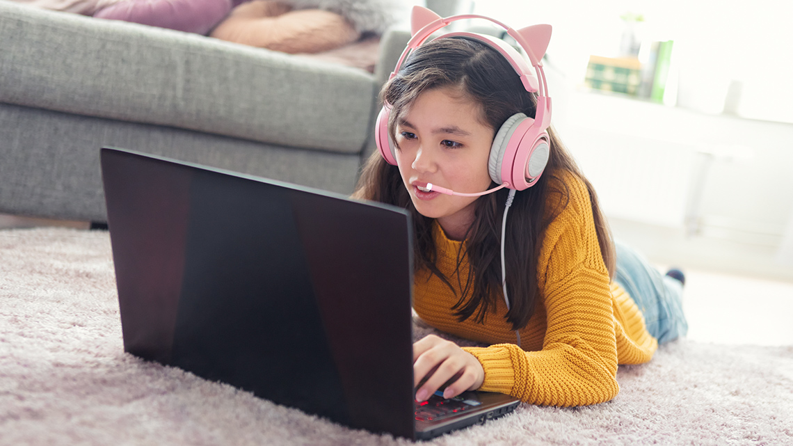 A young person lies on their stomach indoors on a living room carpet wearing a headset and using a laptop to record a podcast.
