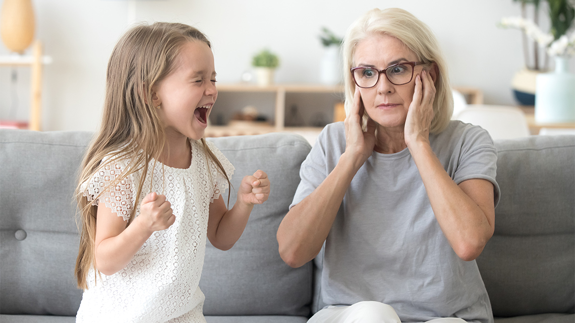 A child screaming with their fists clenched next to an adult that is covering their own ears.