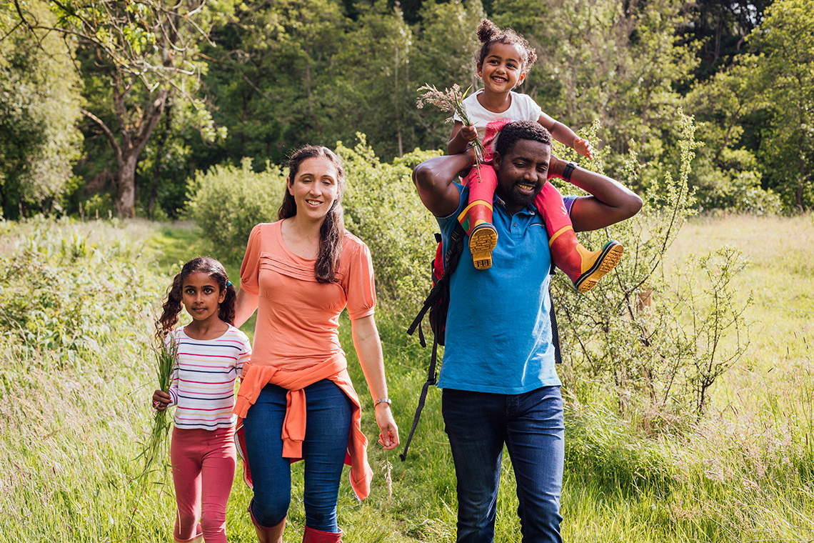 Two adults and two children walk through a field, one child is being held on an adult’s shoulders.