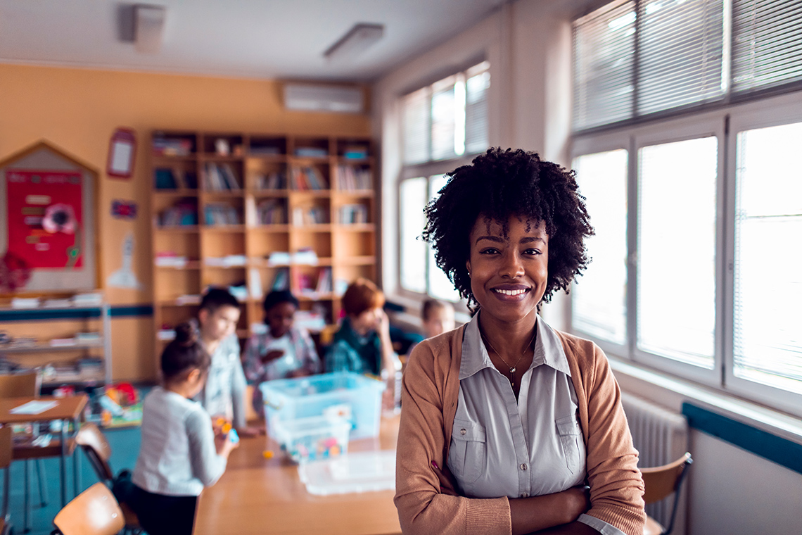 A teacher stands in a classroom with students working together at a table behind them.