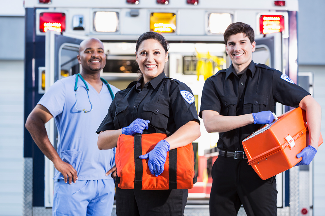 A nurse and two paramedics holding first aid kits in front of an ambulance.