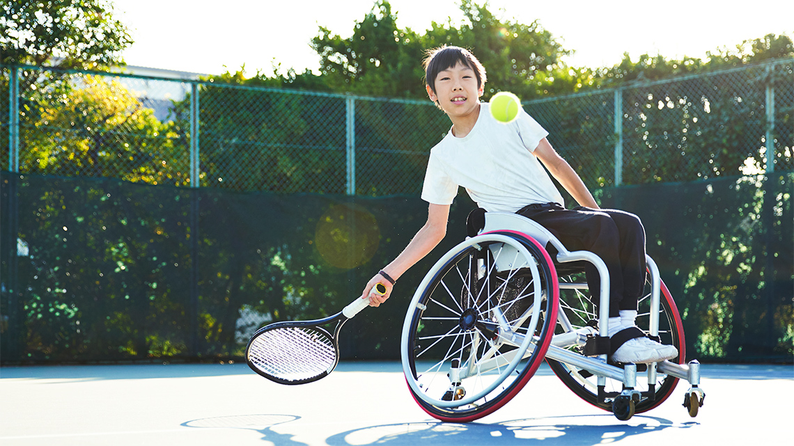 A child who uses a wheelchair is playing tennis and swings their racquet to hit a tennis ball.