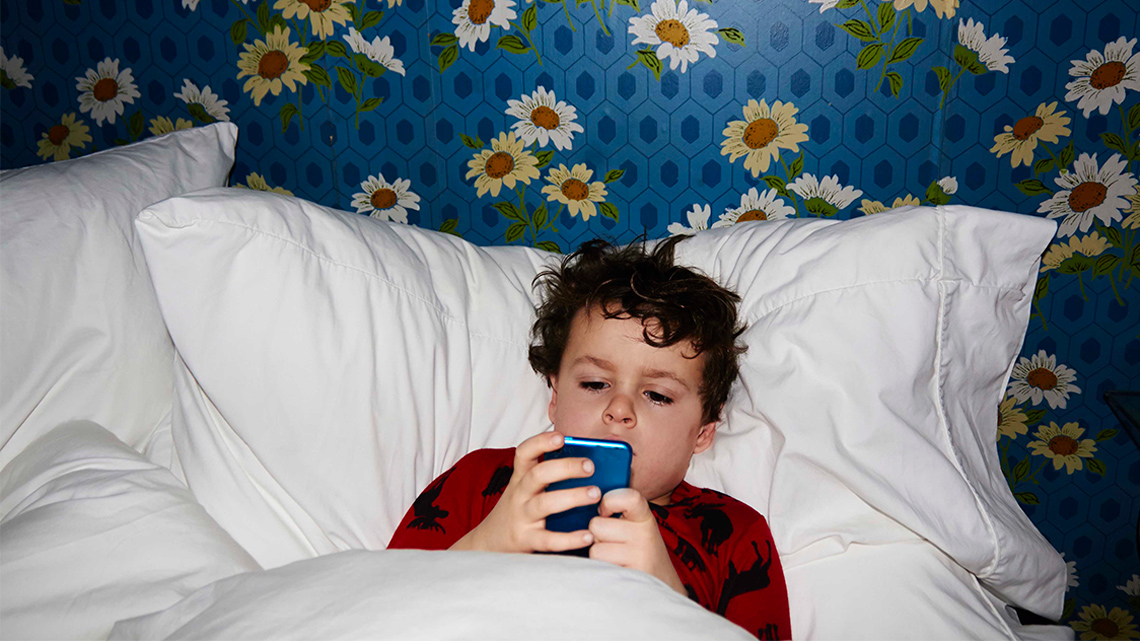 A child lying in bed holding and staring at a cellphone.