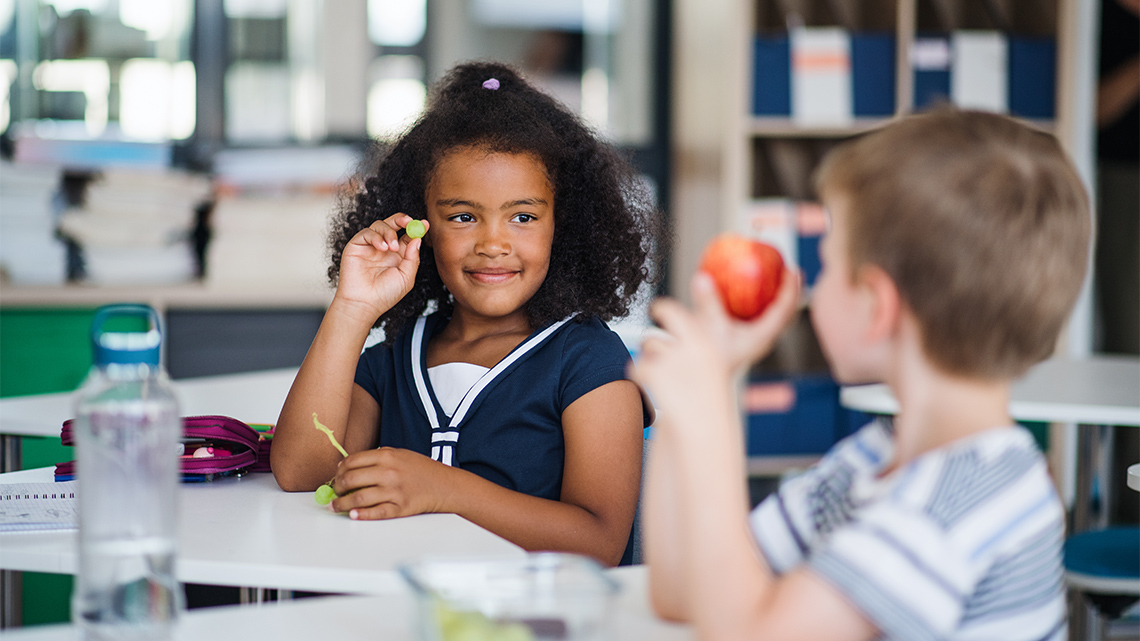 Two students at their desks are showing each other their snacks. One has an apple, one has grapes.