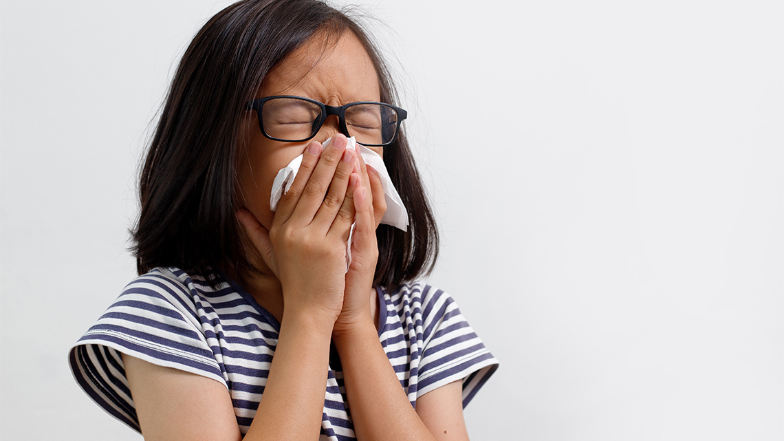 In this image a small child is blowing their nose into a tissue that is covering their whole nose and mouth.