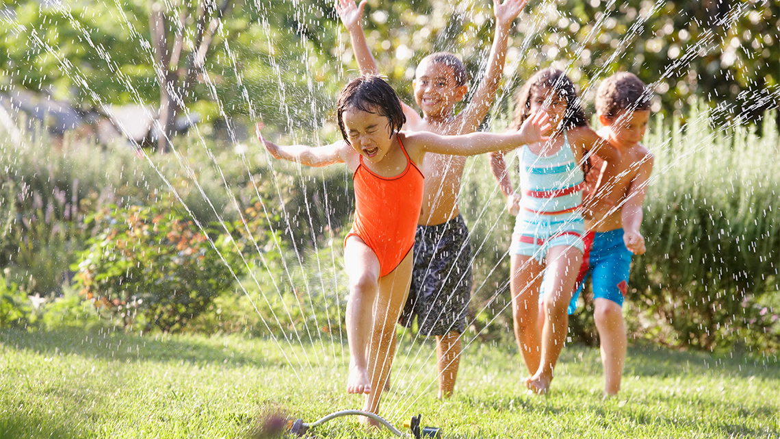 Children in bathing suits are running through a sprinkler on a lawn.