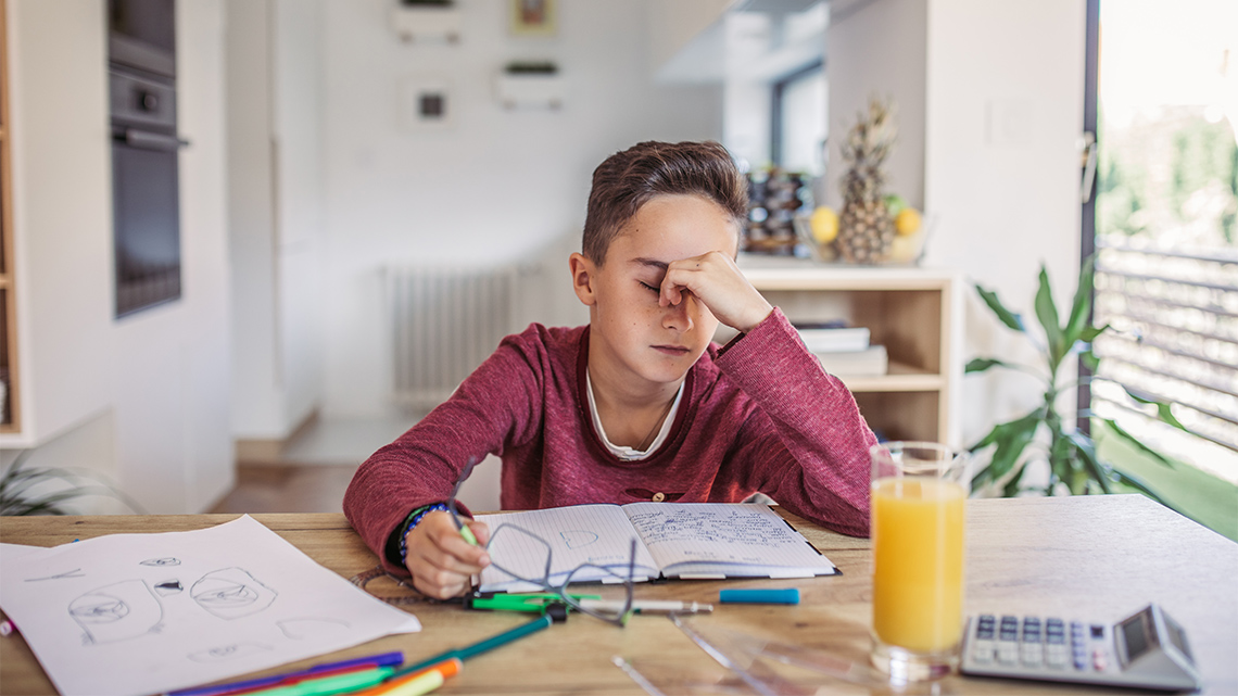 A child is sitting at a desk with some notebooks. Their eyes are closed and they are pinching the bridge of their nose.