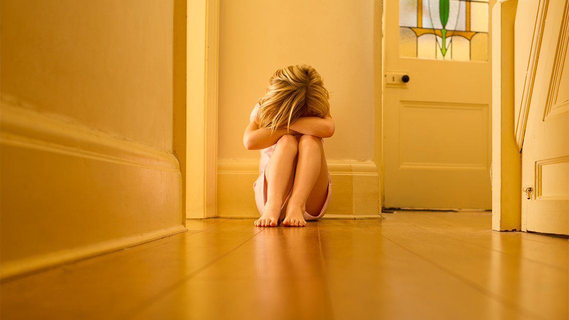 A child crouching in the corner of hallway with their head down and arms crossed.