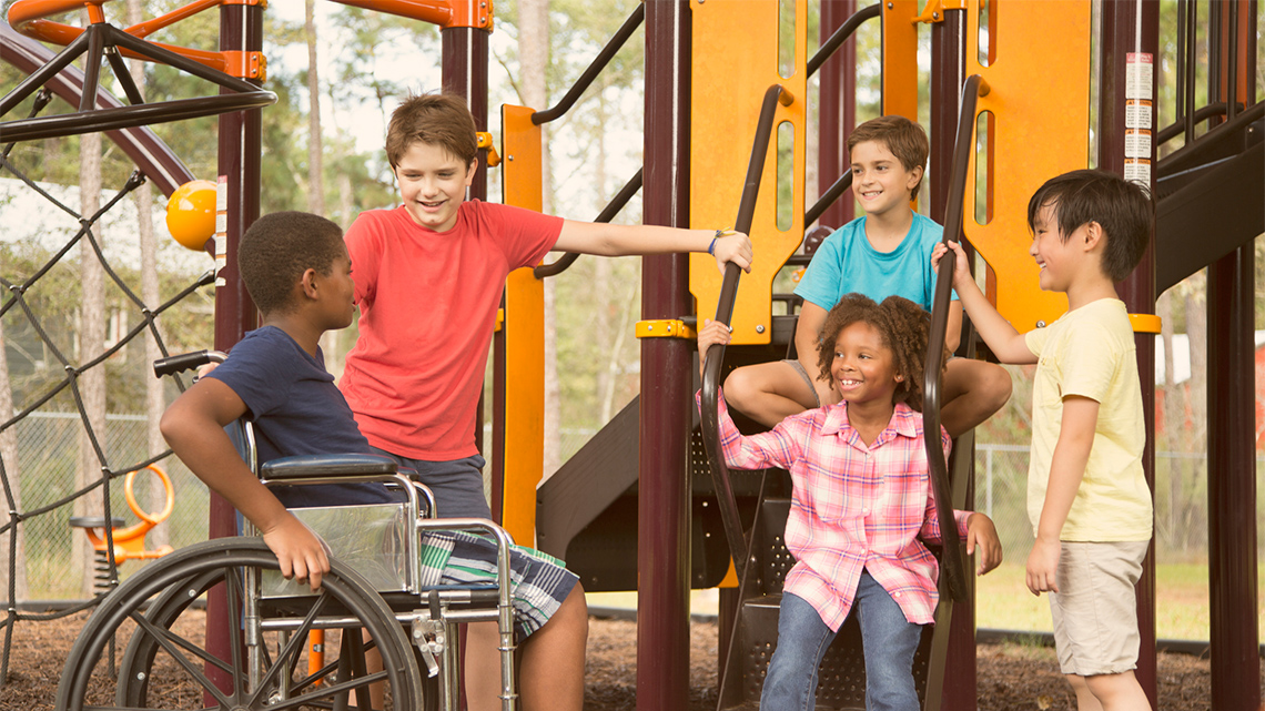 A group of children having fun together at a playground. They are of different ethnicities and one child uses a wheelchair.