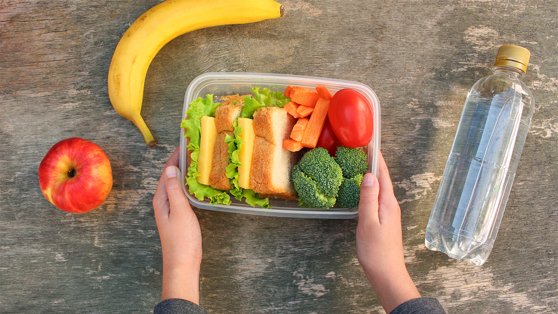 There is a bottle of water, a lunch box with a sandwich and cut up vegetables, a banana, and an apple.