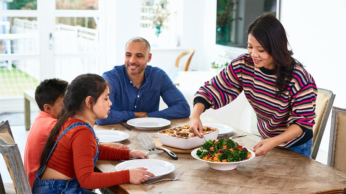 A family eating a meal at the table. There are plates with vegetables and pasta. A parent serves vegetables to a child.